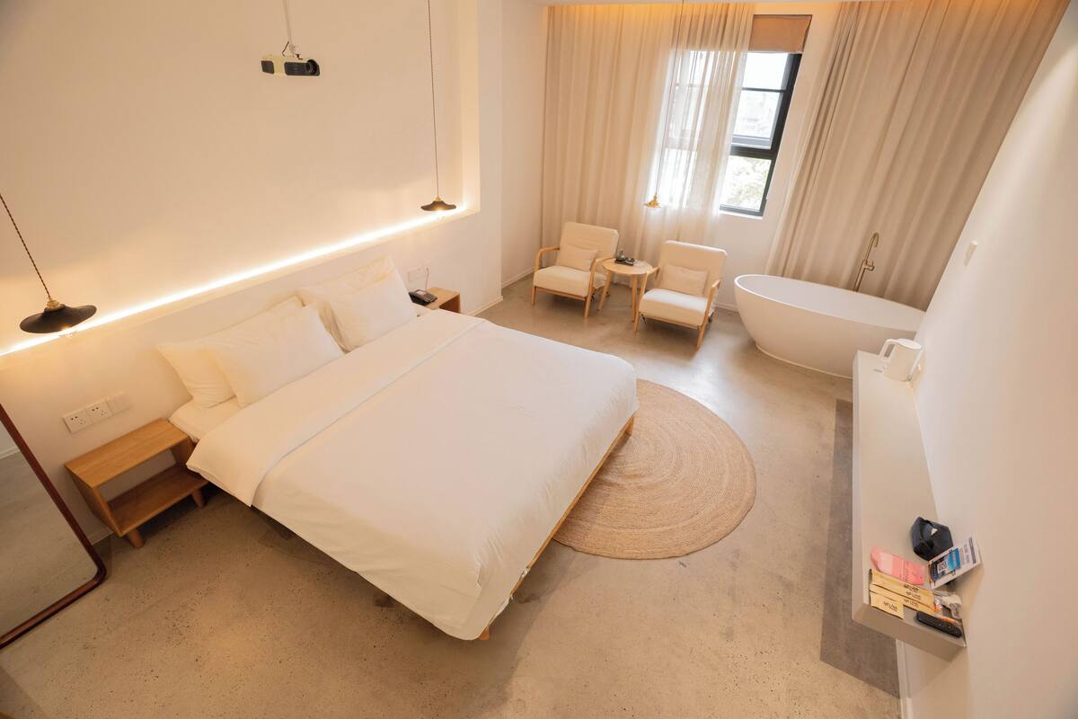 A modern hotel room with a large bed, two armchairs, a small table, a bathtub, and a bedside table with a lamp.