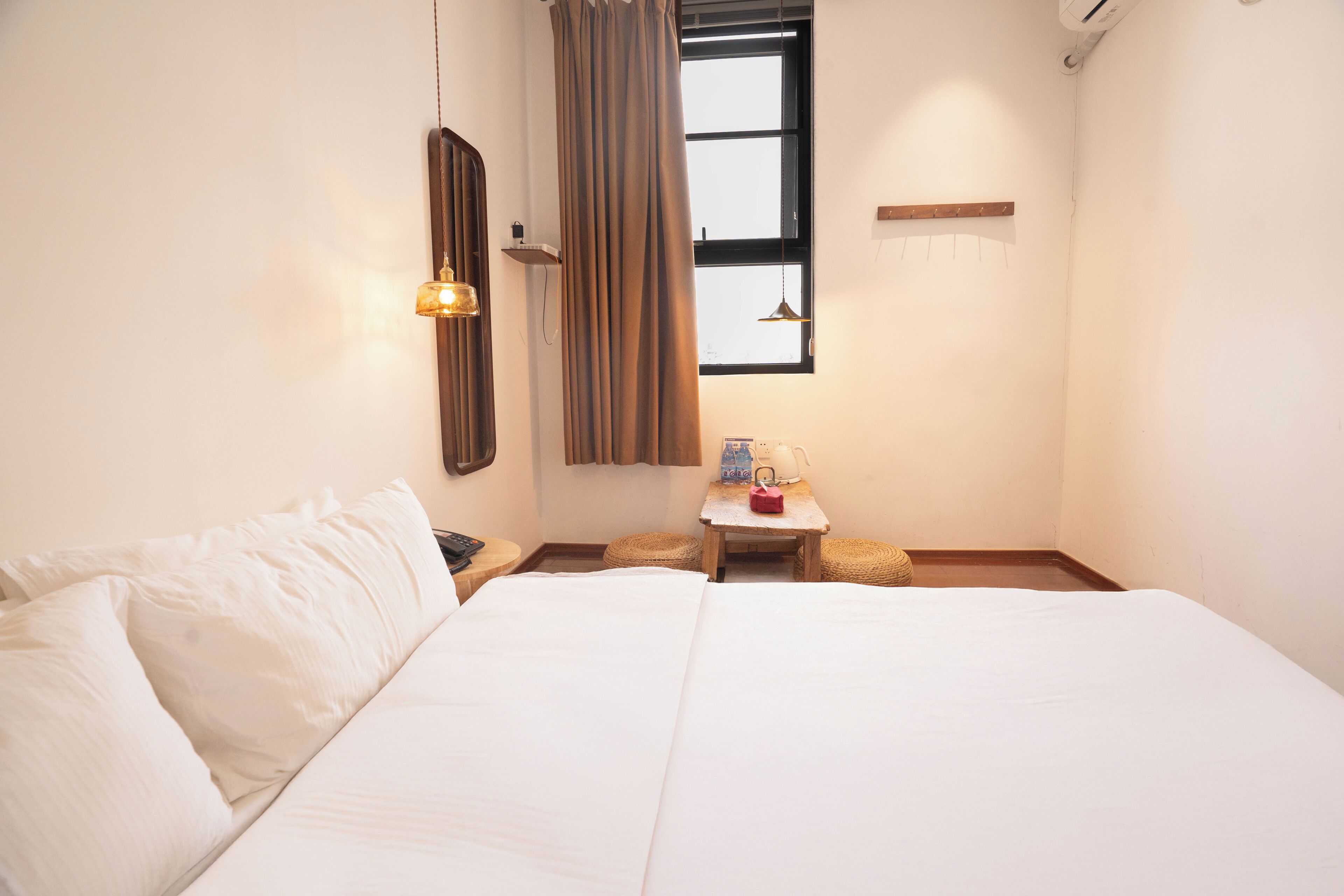 Een hotelkamer met een groot bed, een klein tafeltje met een lamp, een raam met gordijnen, en een plank boven het tafeltje.