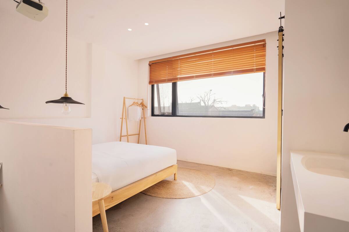 A bedroom with a bed, a bedside table, a hanging light fixture, a window with blinds, and a view of the outside.