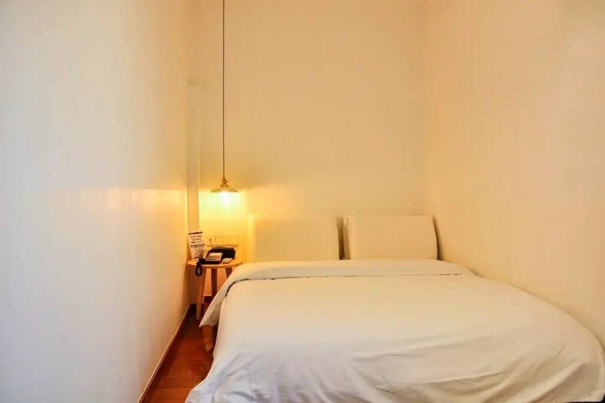 A small, narrow room with a single bed, a bedside table with a lamp, and a telephone.