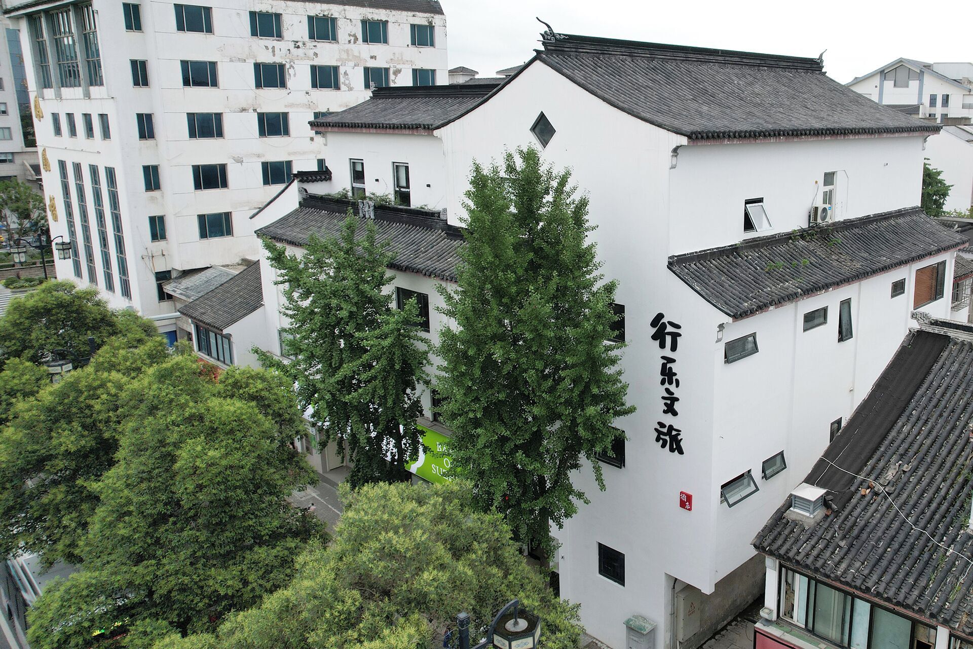 A traditional building with a white facade and black tiled roof, surrounded by greenery and modern buildings.