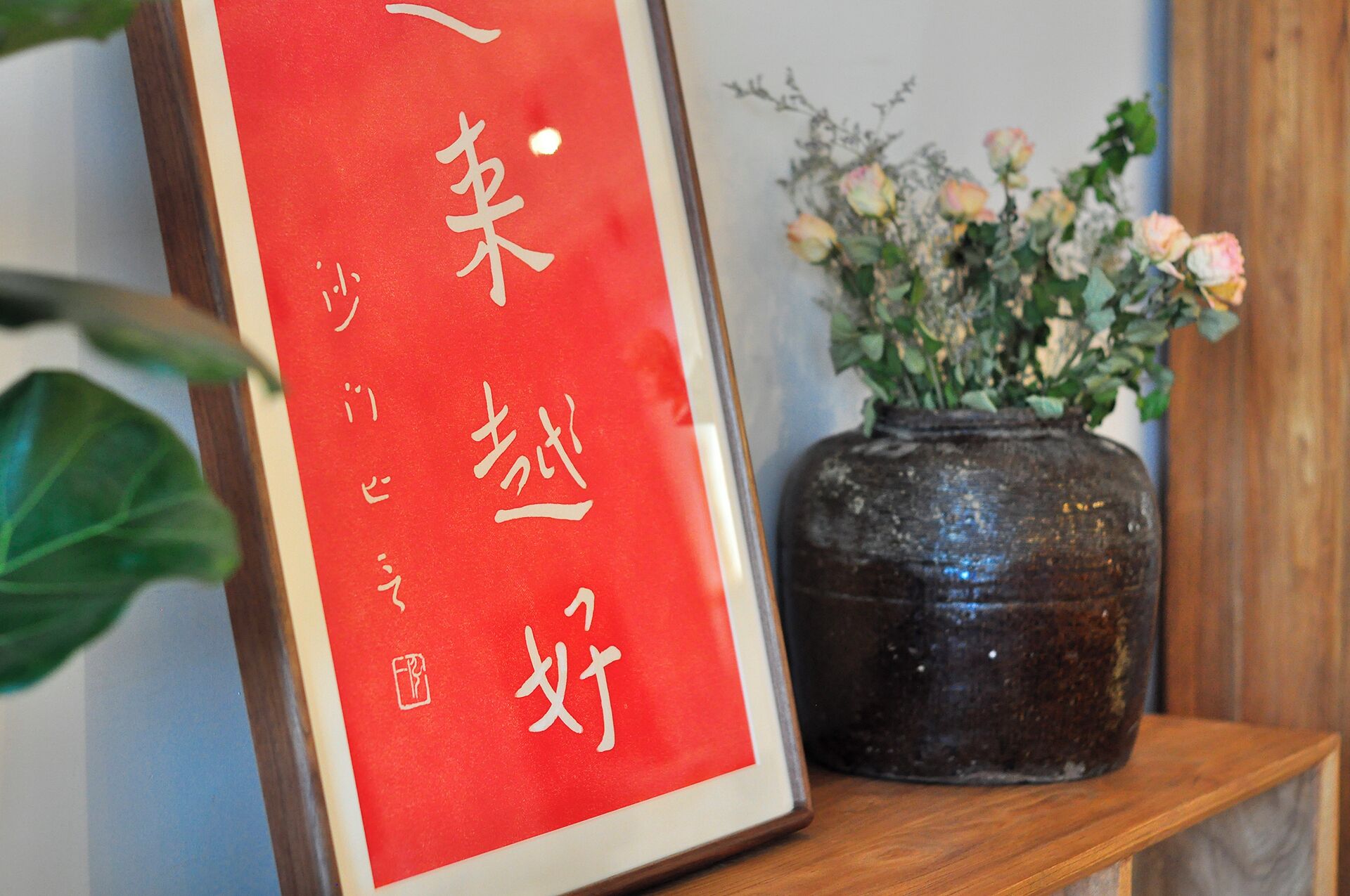 A framed red sign with white calligraphy, a dark ceramic vase with flowers, and a wooden shelf.