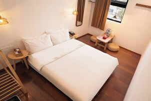 A neatly arranged hotel room with a large bed, a bedside table with a lamp, a small table with a basket, and a window with curtains.