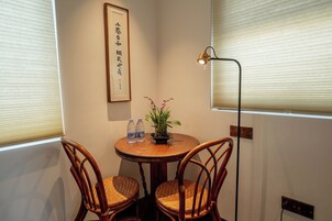 Una habitación pequeña con una mesa redonda, dos sillas de mimbre, un cuadro enmarcado en la pared, una planta en maceta y una lámpara de pie.