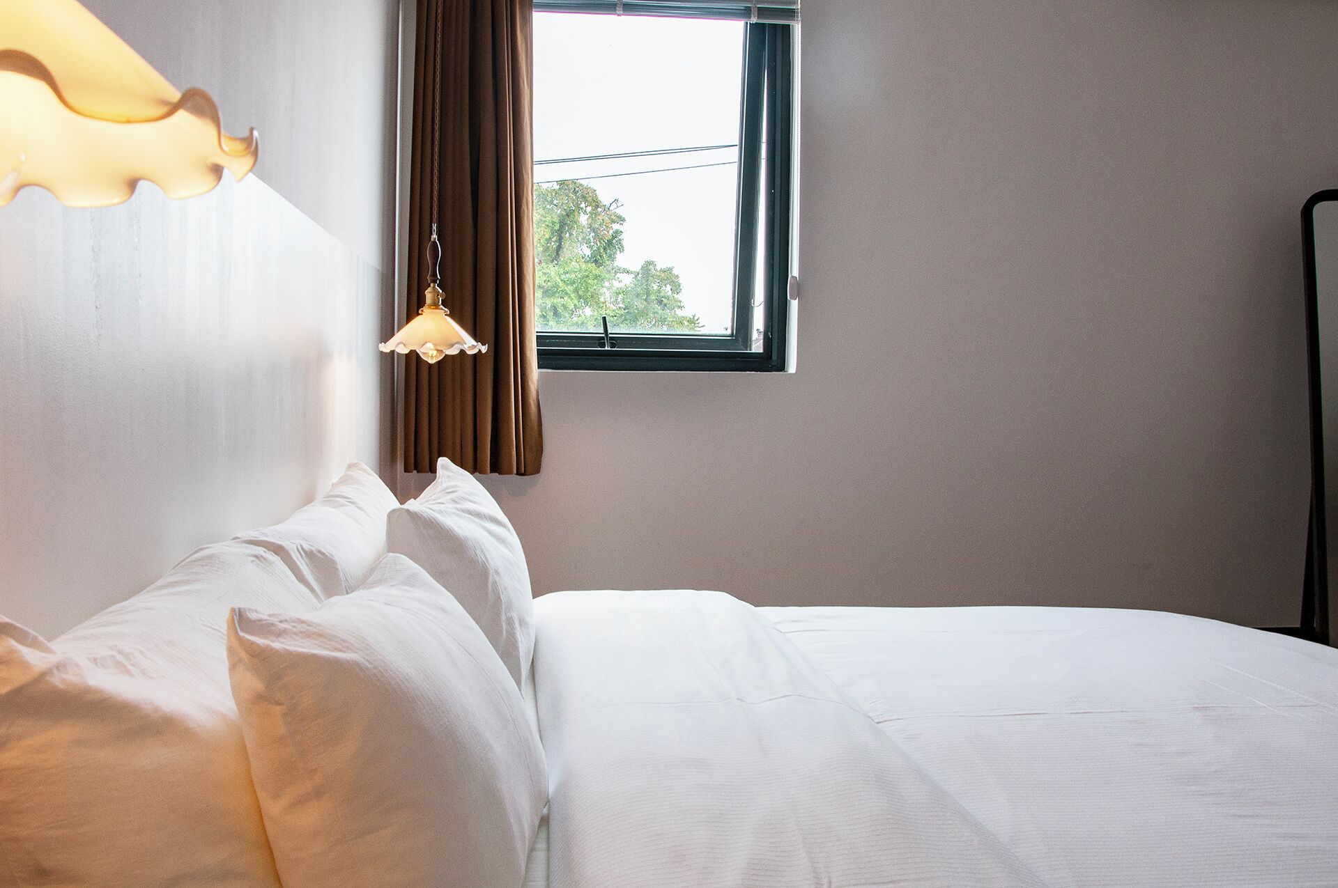 A hotel room with a bed, white bedding, a hanging lamp, a window with curtains, and a view of trees outside.
