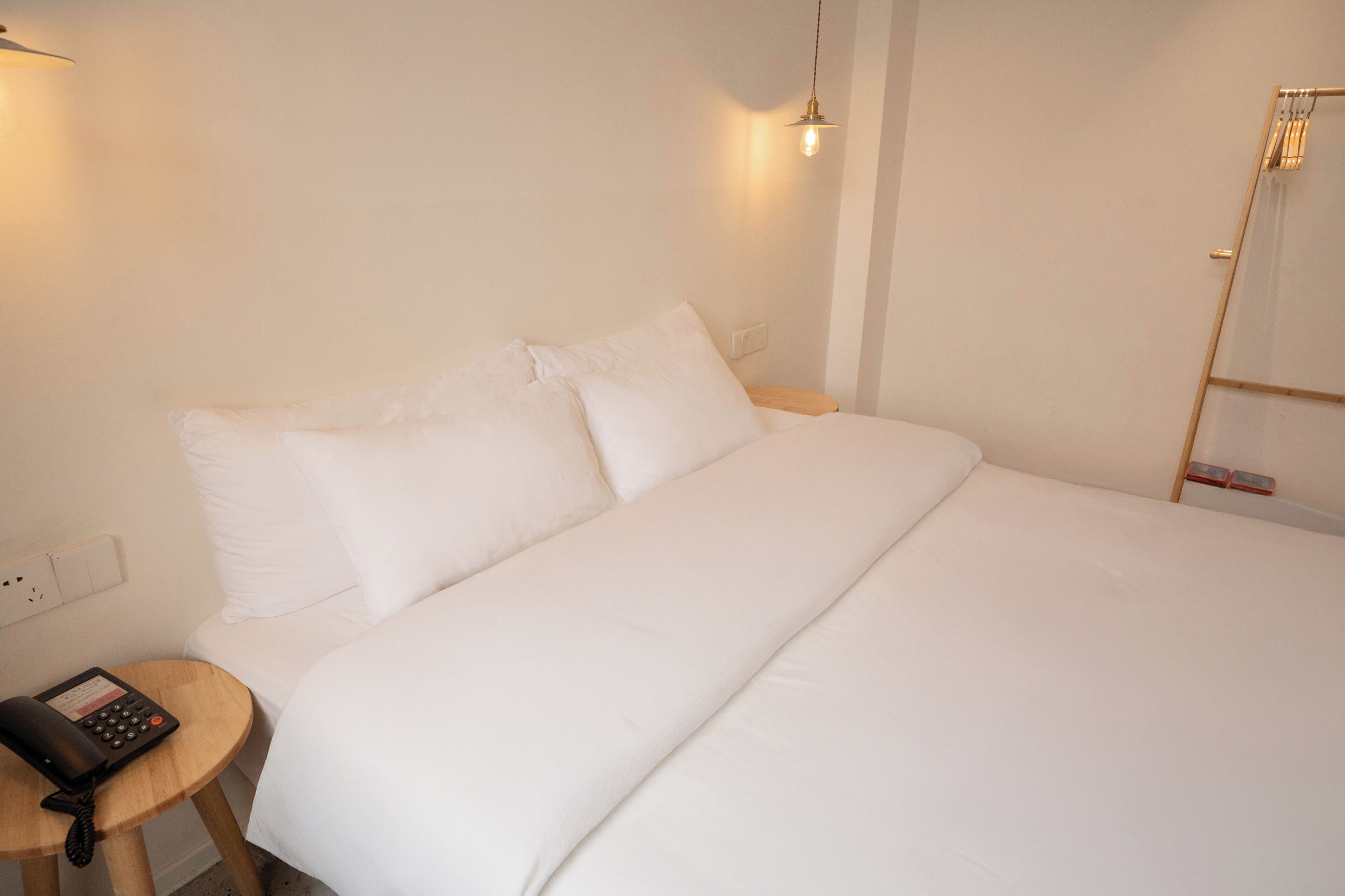 A neatly made bed with white linens, a bedside table with a telephone, and a wall-mounted light fixture.
