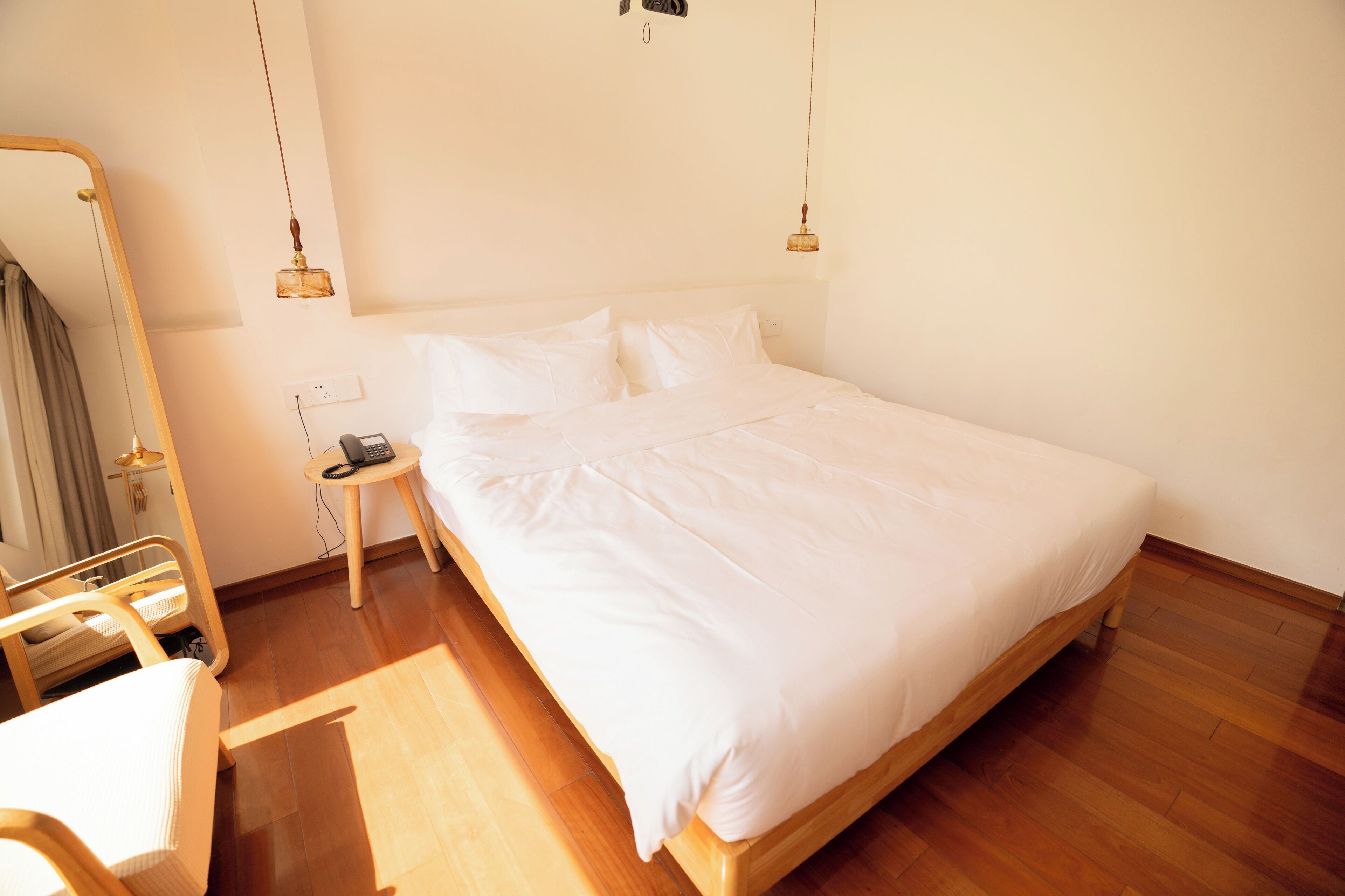 Una cama bien hecha con ropa blanca, piso de madera, una mesita de noche con lámpara y un espejo en la pared.