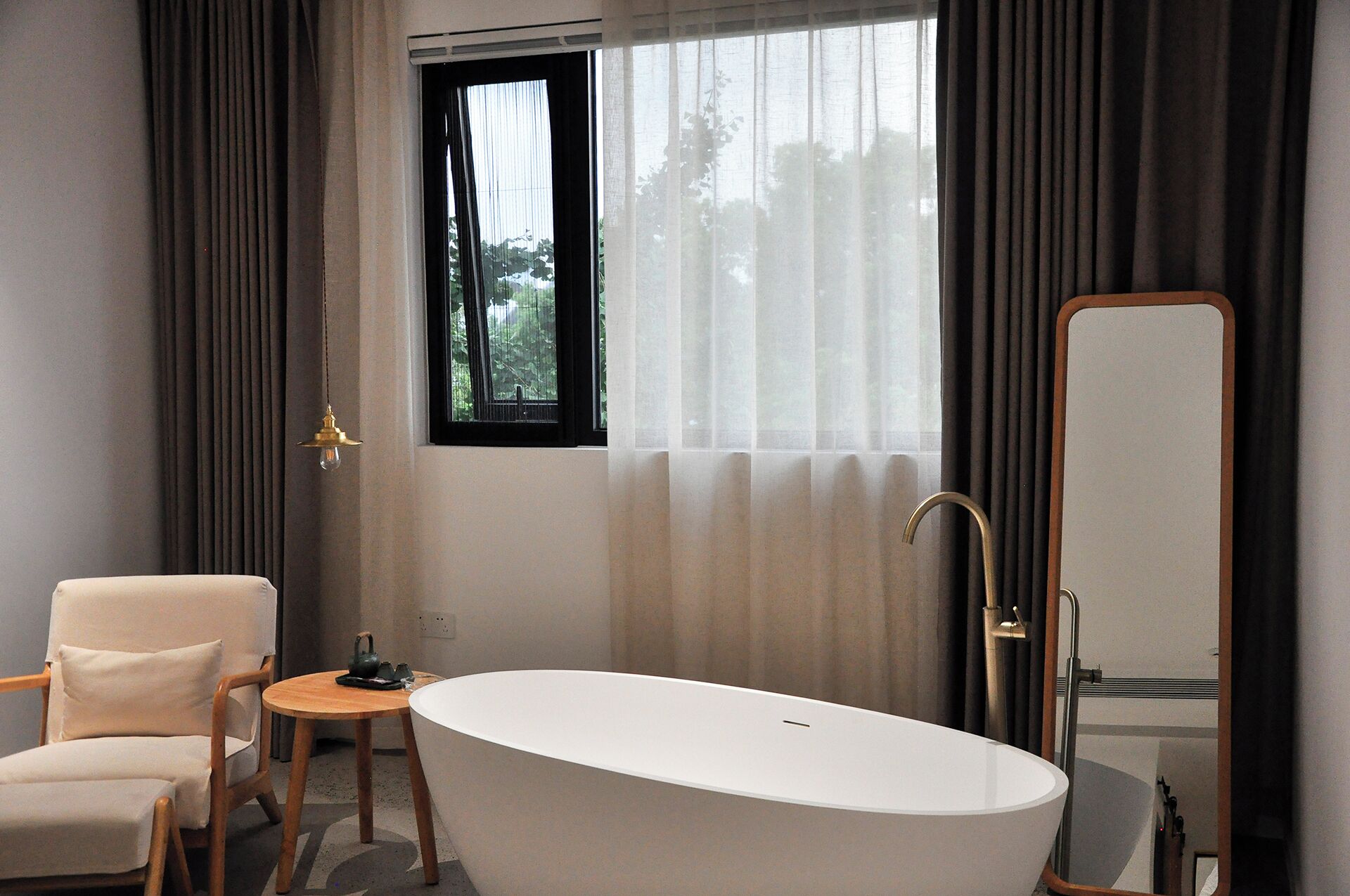 A modern bathroom with a freestanding bathtub, a wooden chair, a small round table, and a large mirror.