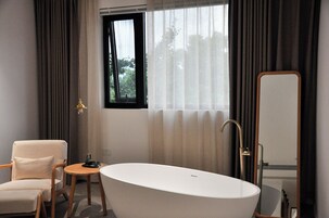 A modern bathroom with a freestanding bathtub, a wooden chair, a small round table, and a large mirror.