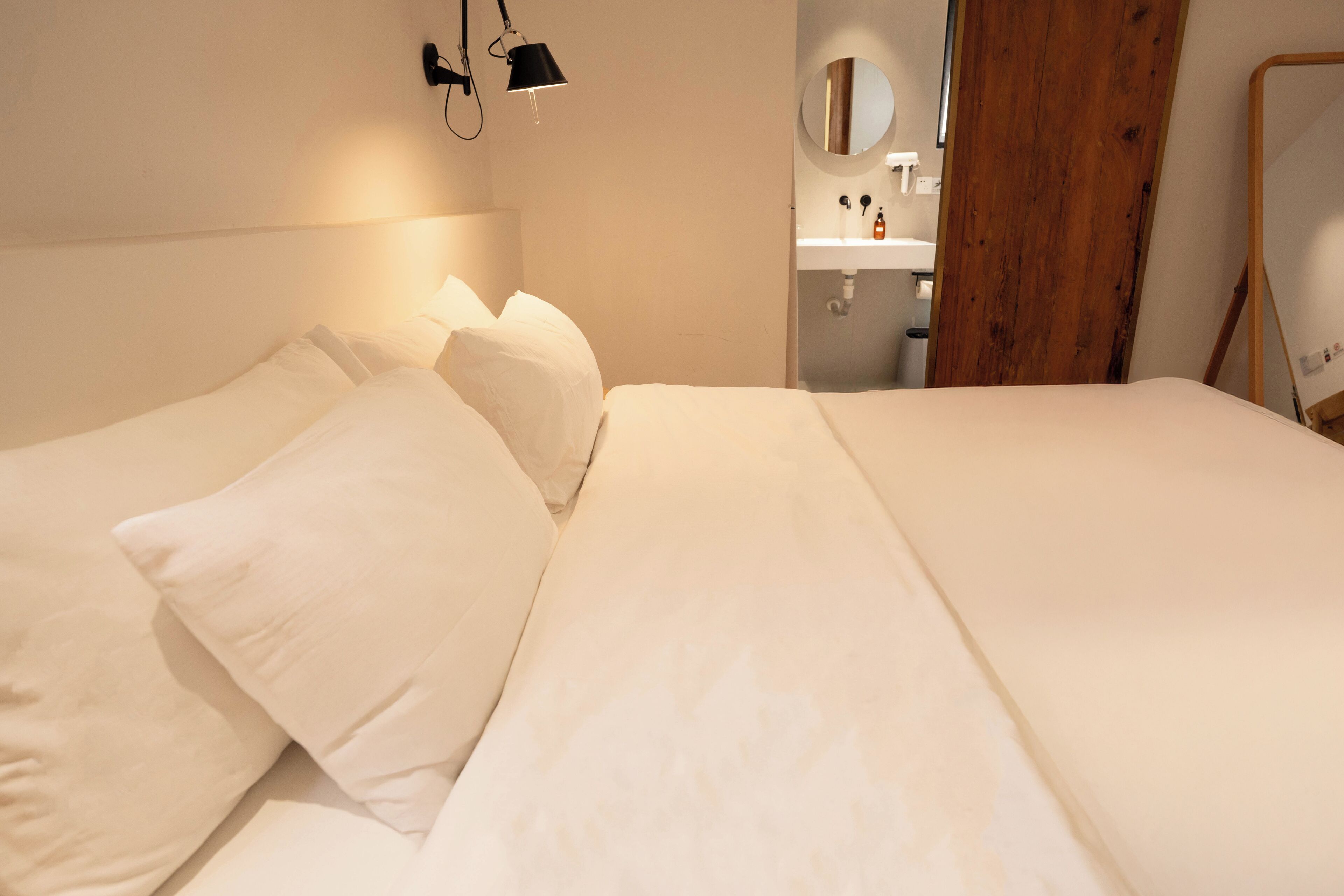 A neatly made bed with white linens and pillows, a bedside lamp, a wooden door, and a small bathroom with a mirror and sink.