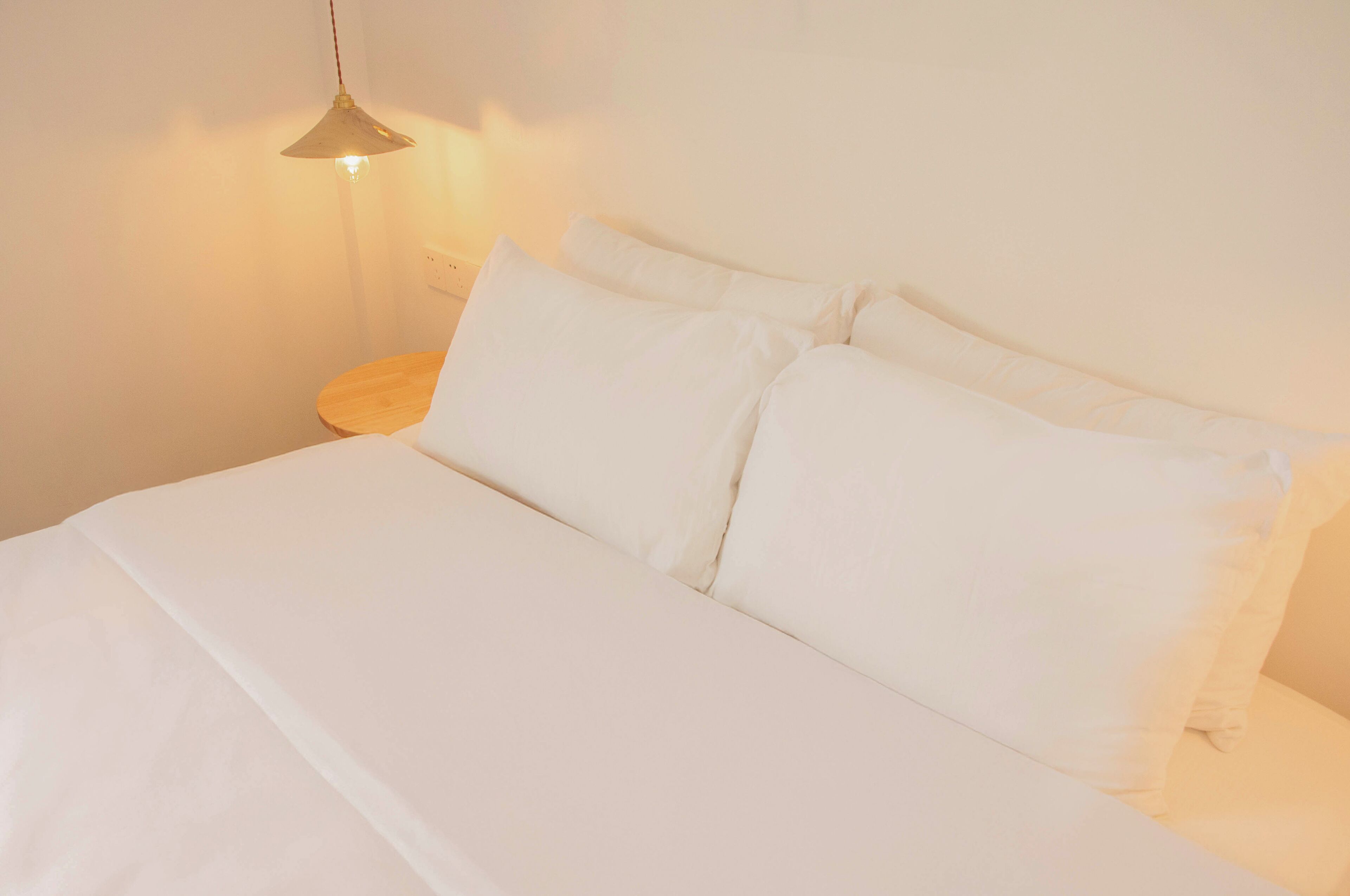 Una cama bien tendida con sábanas y almohadas blancas, una mesita de noche con lámpara, y una pared lisa.