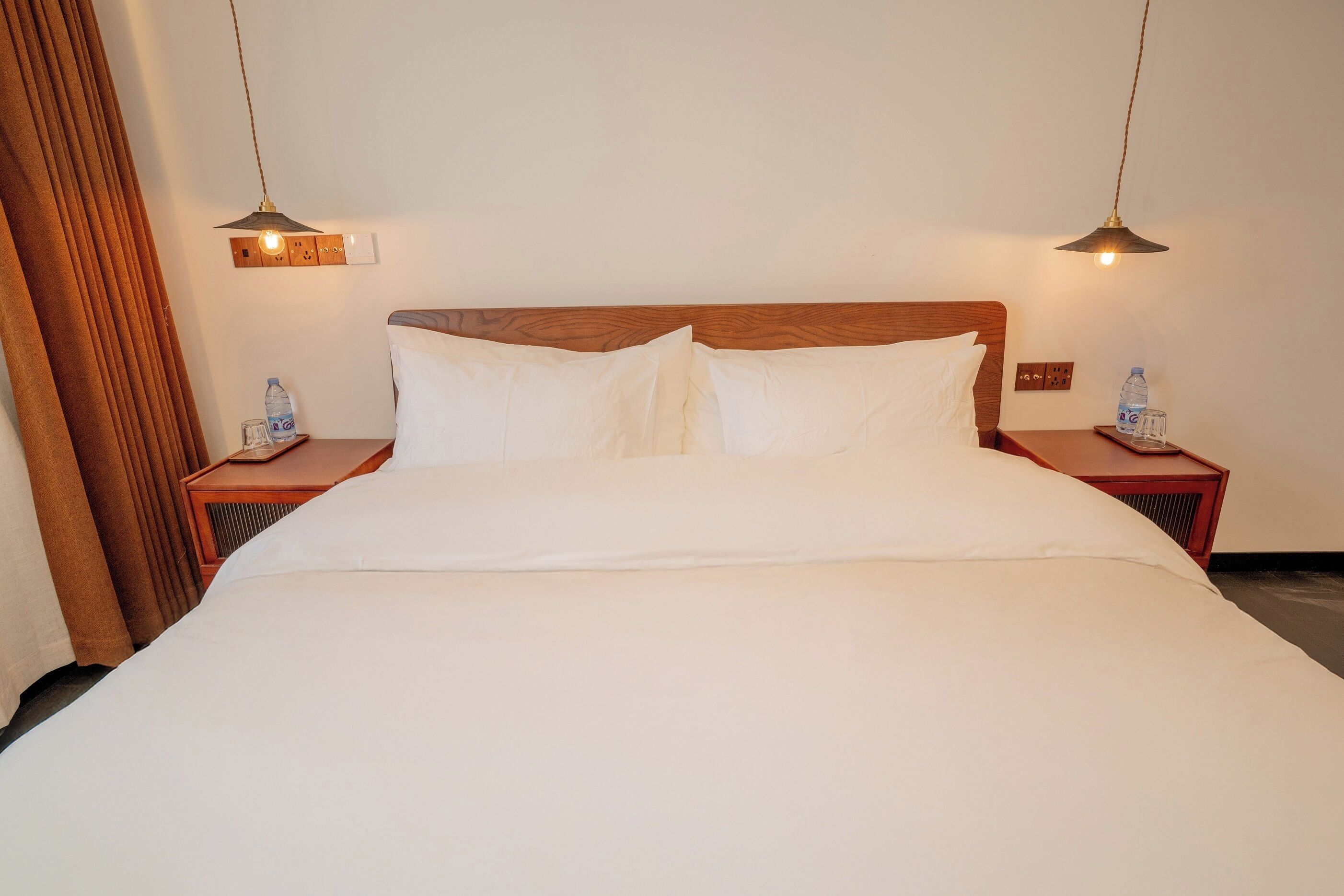 A neatly made bed with white linens, two bedside tables with water bottles, and pendant lights above.