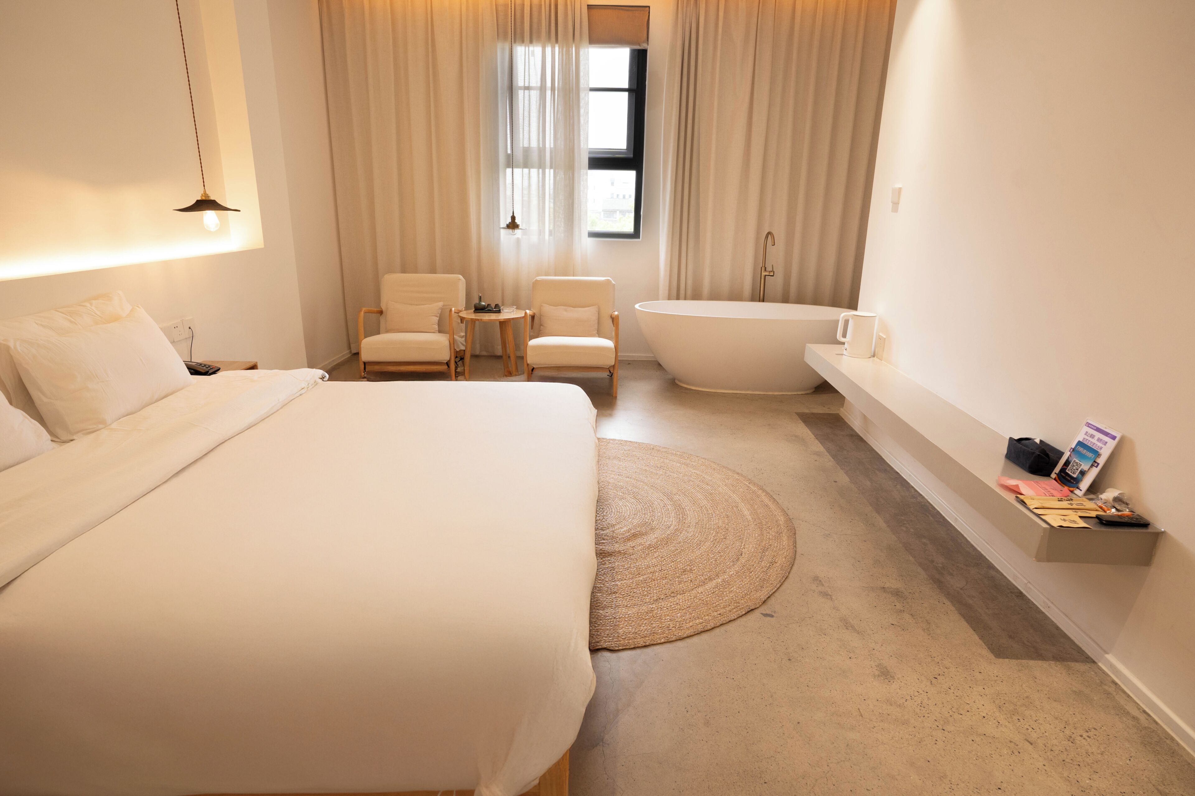 Una habitación de hotel con una cama grande, dos sillas, una mesita, una bañera y un estante con objetos.