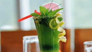 Un vaso alto con una bebida verde, adornado con hojas de menta, rodajas de limón y una pajita roja.