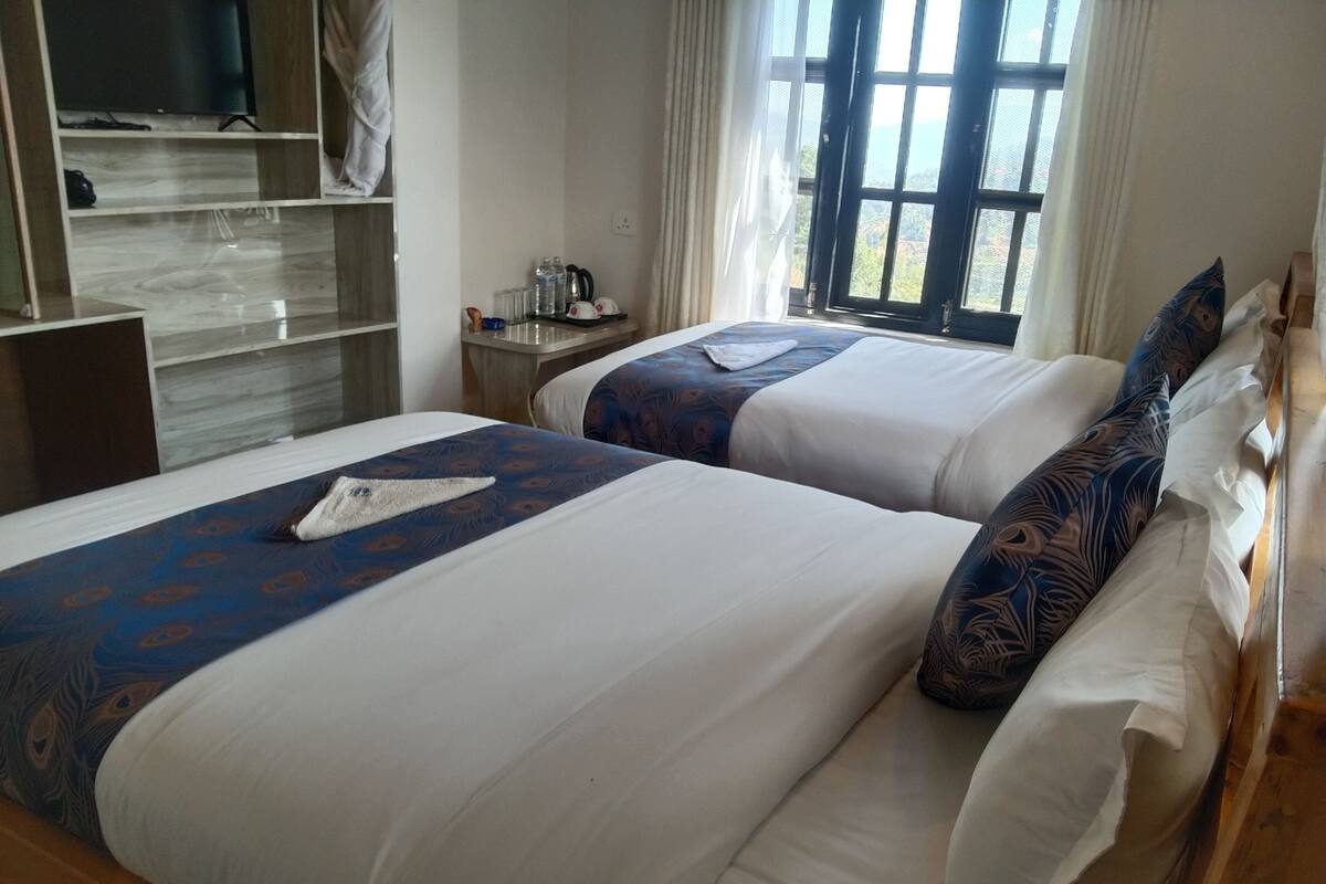 Two beds with white and blue bedding, a wooden desk, a television, and a window with curtains.