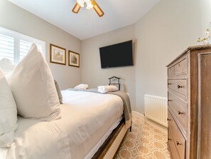 Een net opgemaakt bed met kussens, een dressoir, een televisie aan de muur en ingelijste foto's.
