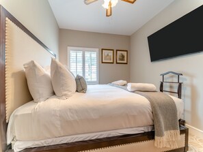 A bedroom with a bed, pillows, a bedside table, a TV, and a window with blinds.
