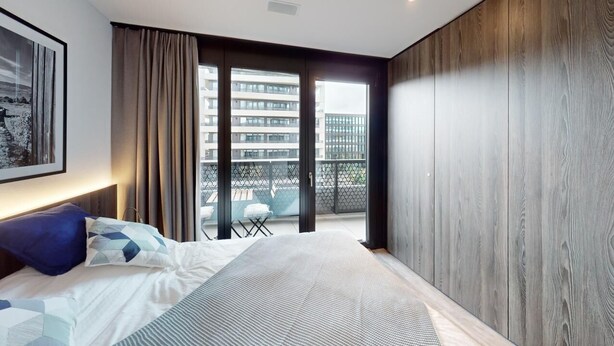 A modern bedroom with a large bed, a view of the city through floor-to-ceiling windows, and a balcony with outdoor furniture.