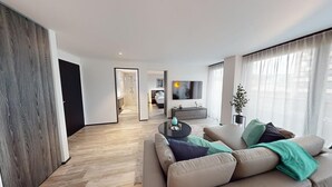 A modern living room with a sofa, a coffee table, and a TV. There is a door leading to a bathroom and a hallway.