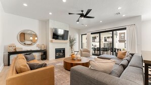 A modern living room with a fireplace, a flat-screen TV mounted on the wall, a ceiling fan, and a large window with curtains. - Modern Oasis at Desert Color (by Ember Stays) (St. George)