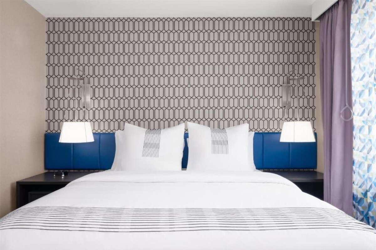 A hotel room with a patterned wallpaper, a large bed with a striped bedspread, two bedside tables with lamps, and a headboard with a blue accent.