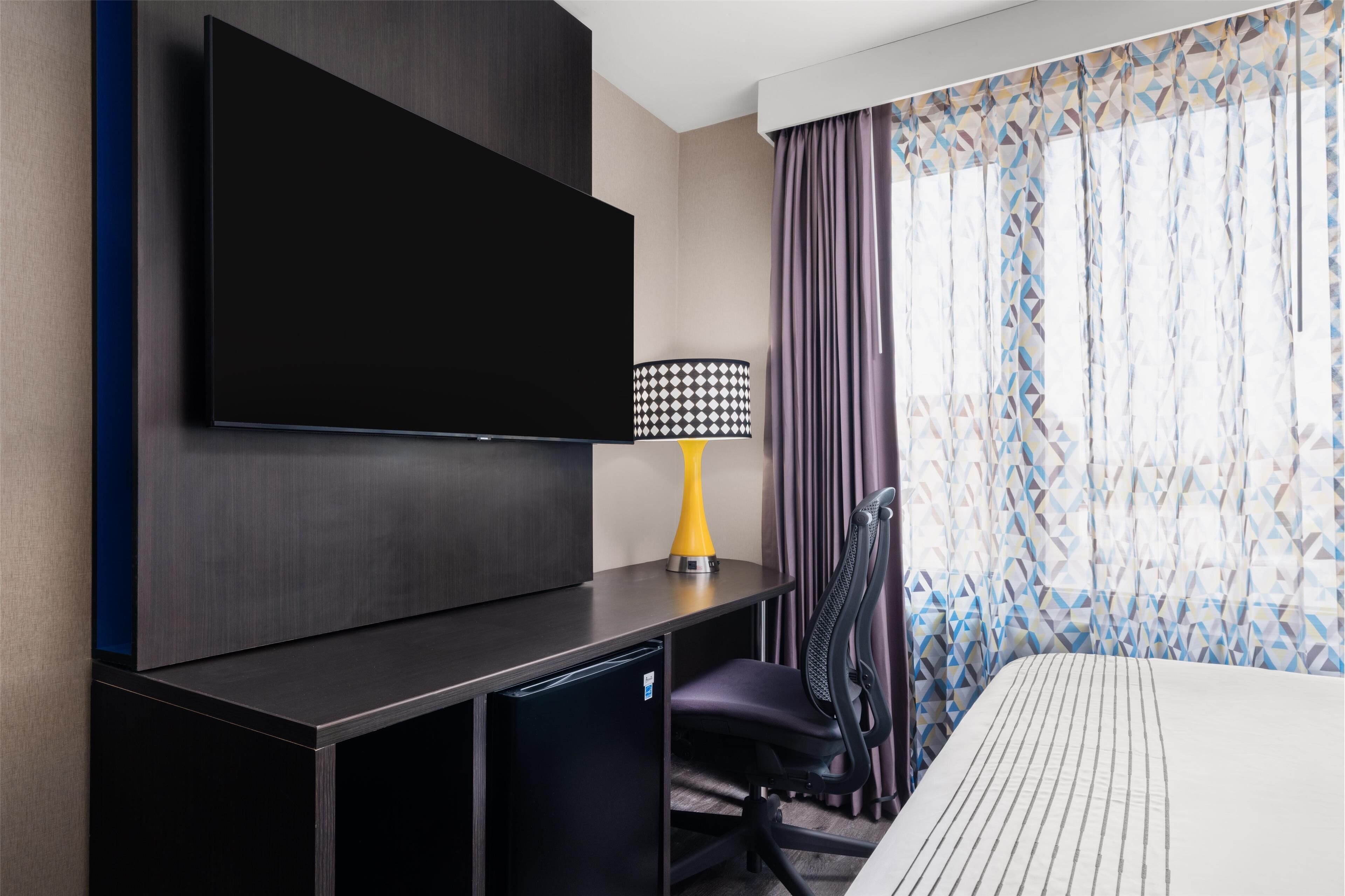 Une chambre d’hôtel équipée d’un téléviseur à écran plat fixé au mur, d’un bureau avec une lampe, d’une chaise et d’un lit recouvert d’une courtepointe à rayures.