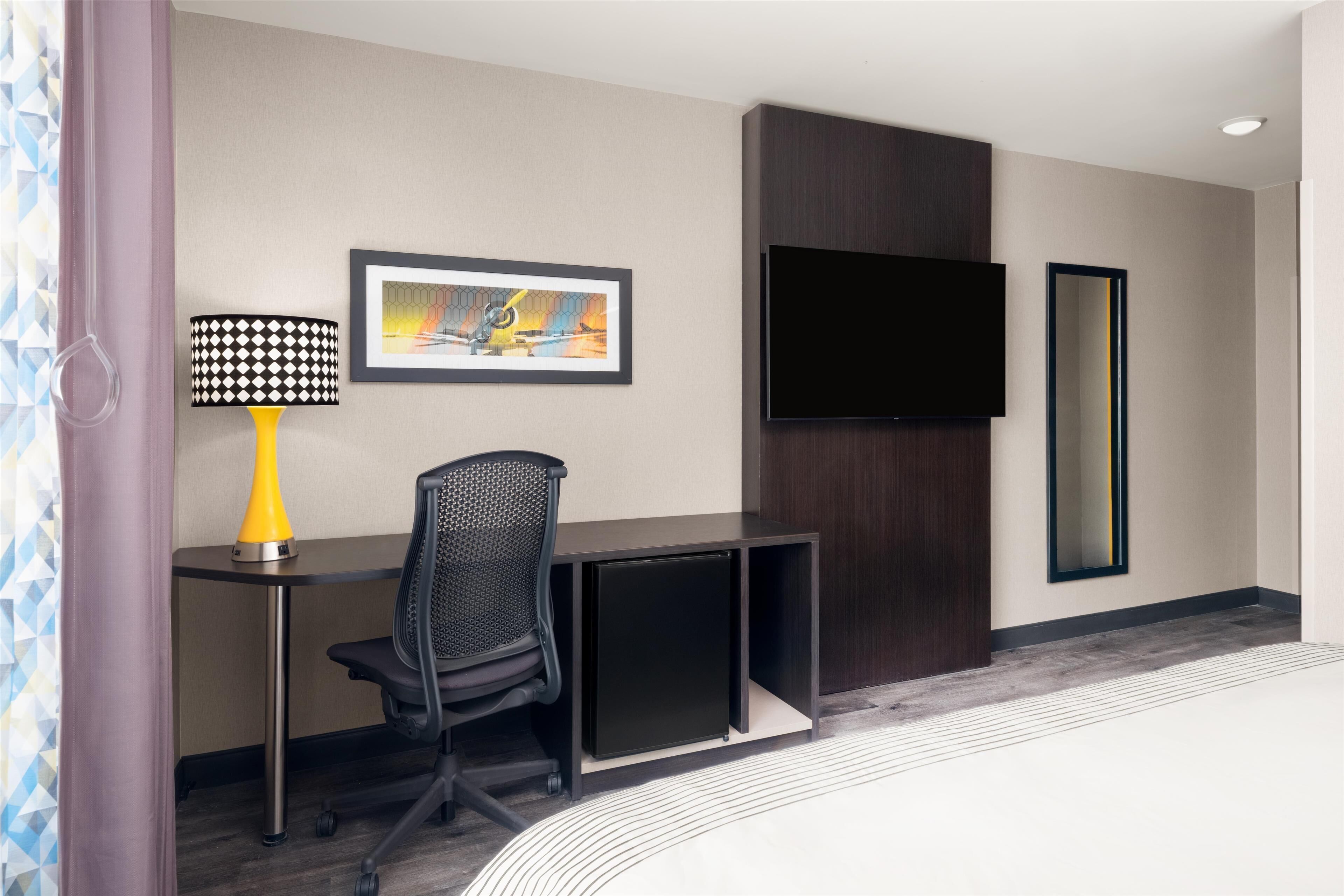 Habitación de hotel con escritorio, silla, lámpara, televisor y un espejo grande.