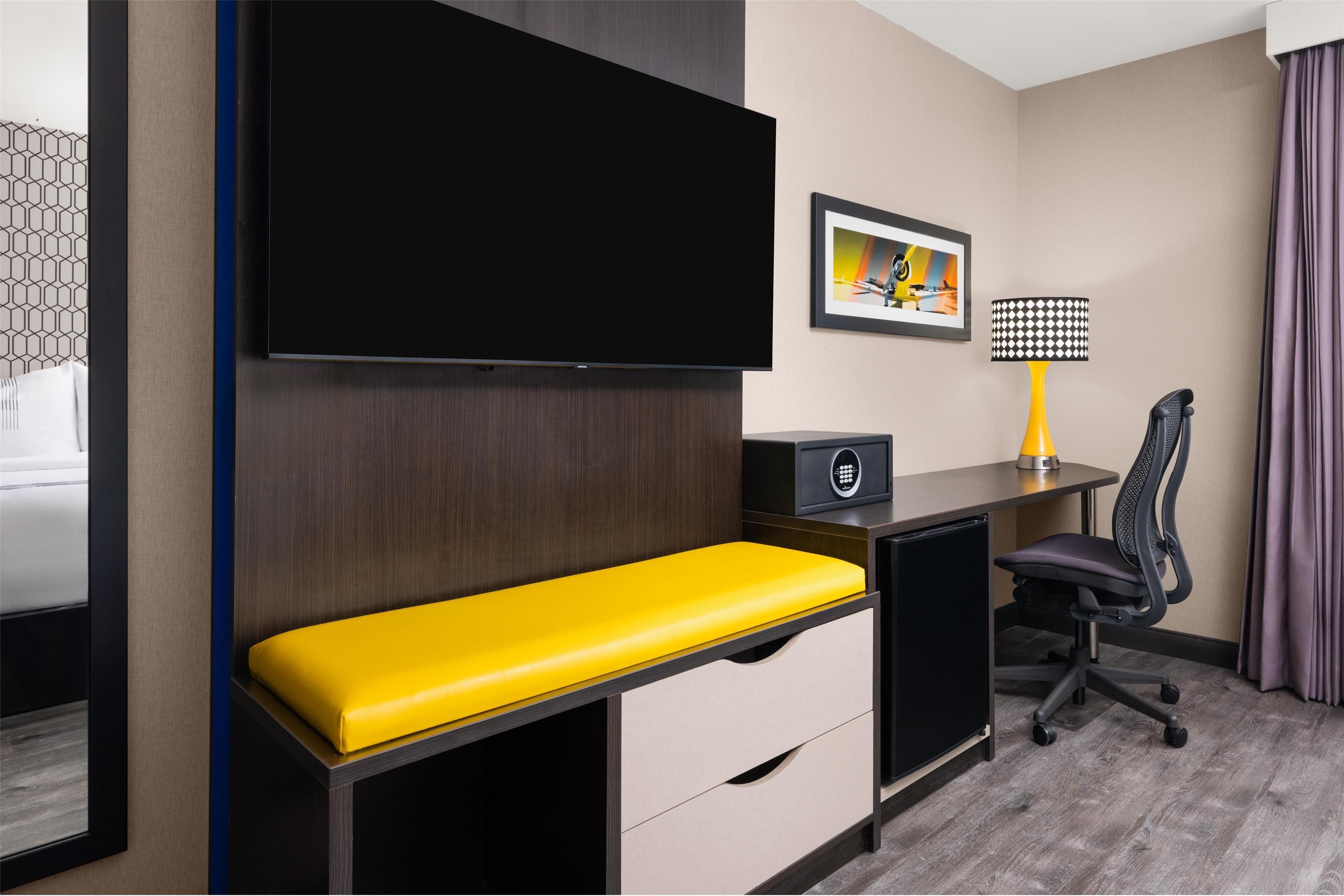 A hotel room with a flat-screen TV mounted on the wall, a desk with a lamp, a chair, and a bench.