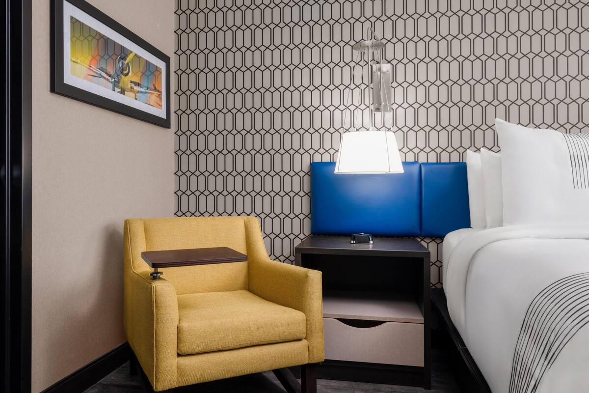 Une chambre d’hôtel comprenant un lit, une table de chevet, une lampe, une chaise et un mur orné d’un motif géométrique.