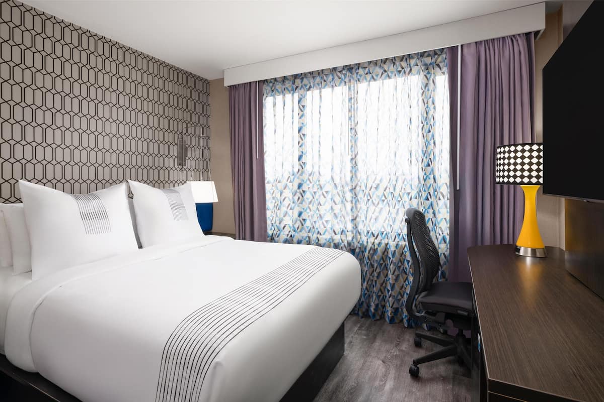 Une chambre d’hôtel avec un grand lit, un bureau avec une lampe, une chaise et une fenêtre avec des rideaux à motifs.