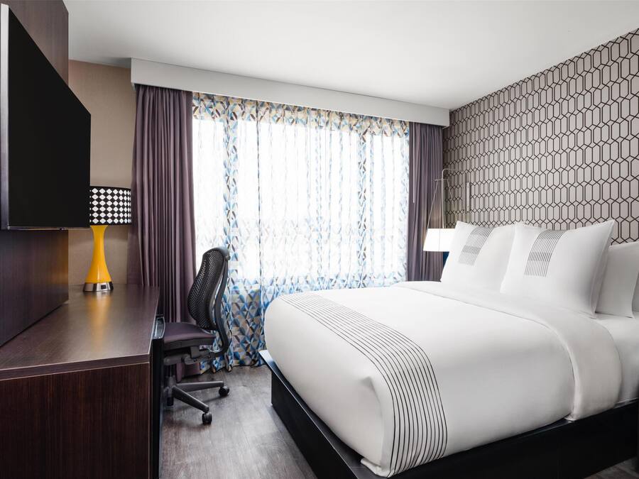 A hotel room with a large bed, a desk with a chair, a TV, and a patterned wall.