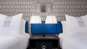 Habitación de hotel de diseño moderno, con un muro de patrón geométrico, un cabecero azul, dos camas blancas con almohadas a juego, una mesita de noche con lámpara y una caja fuerte.
