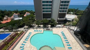 Une piscine sur le toit, dotée de chaises longues, offrant une vue sur l’océan et avec un grand bâtiment en arrière-plan.