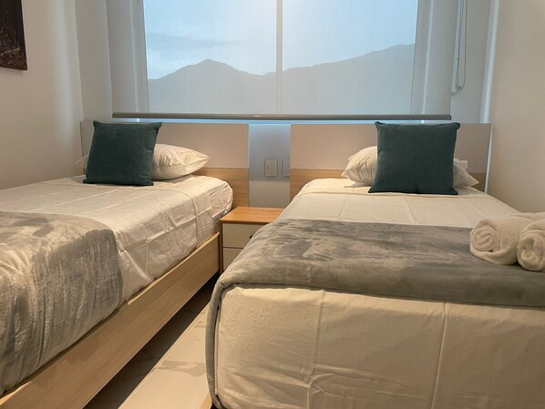 Habitación de hotel con dos camas, cada una con almohada y manta. Se ve una ventana con vista a montañas.