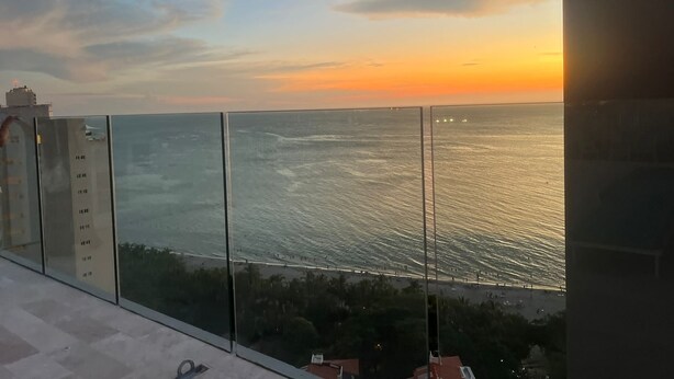 Une vue sur le coucher de soleil depuis le balcon d’un immeuble de grande hauteur, donnant sur une plage sous un ciel dégagé.