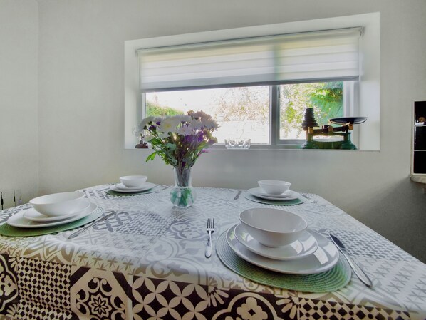A dining table set with white plates, bowls, and cutlery, a vase with flowers, and a window with blinds.