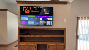 A flat-screen TV mounted on a wall displaying various streaming services. Below the TV, there is a wooden entertainment center with two doors and a shelf. - Charming remodeled 2-bedroom furnished home in safe, delightful Menomonee Falls (Menomonee Falls)