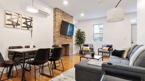 A modern living room with a brick fireplace, a dining area with black chairs, a grey sofa, and a large window.