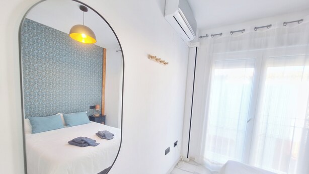 A modern hotel room with a large mirror, a bed with white linens, a bedside table with a lamp, and a window with sheer curtains. - THE PORT. E151A VacaHome Management (El Puerto de Santa María)