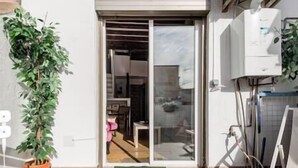 Une entrée moderne avec une porte coulissante en verre, une plante en pot et un espace intérieur visible, où l’on aperçoit un fauteuil rose.