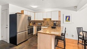 A modern kitchen with stainless steel appliances, a granite countertop, and a small dining area with two chairs. - Gold-rated 2 Bedroom Condo at the Base of Aspen Mountain (Aspen)