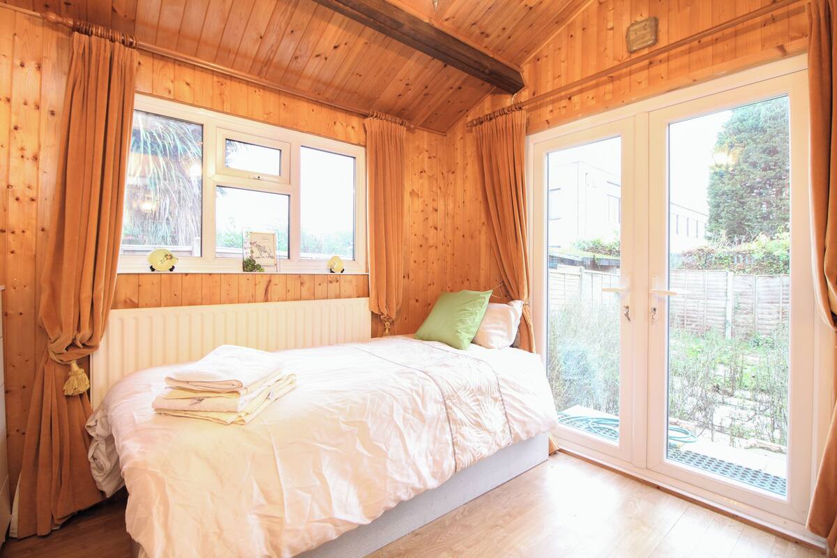 A wooden room with a bed, two windows, and a view of the outside.