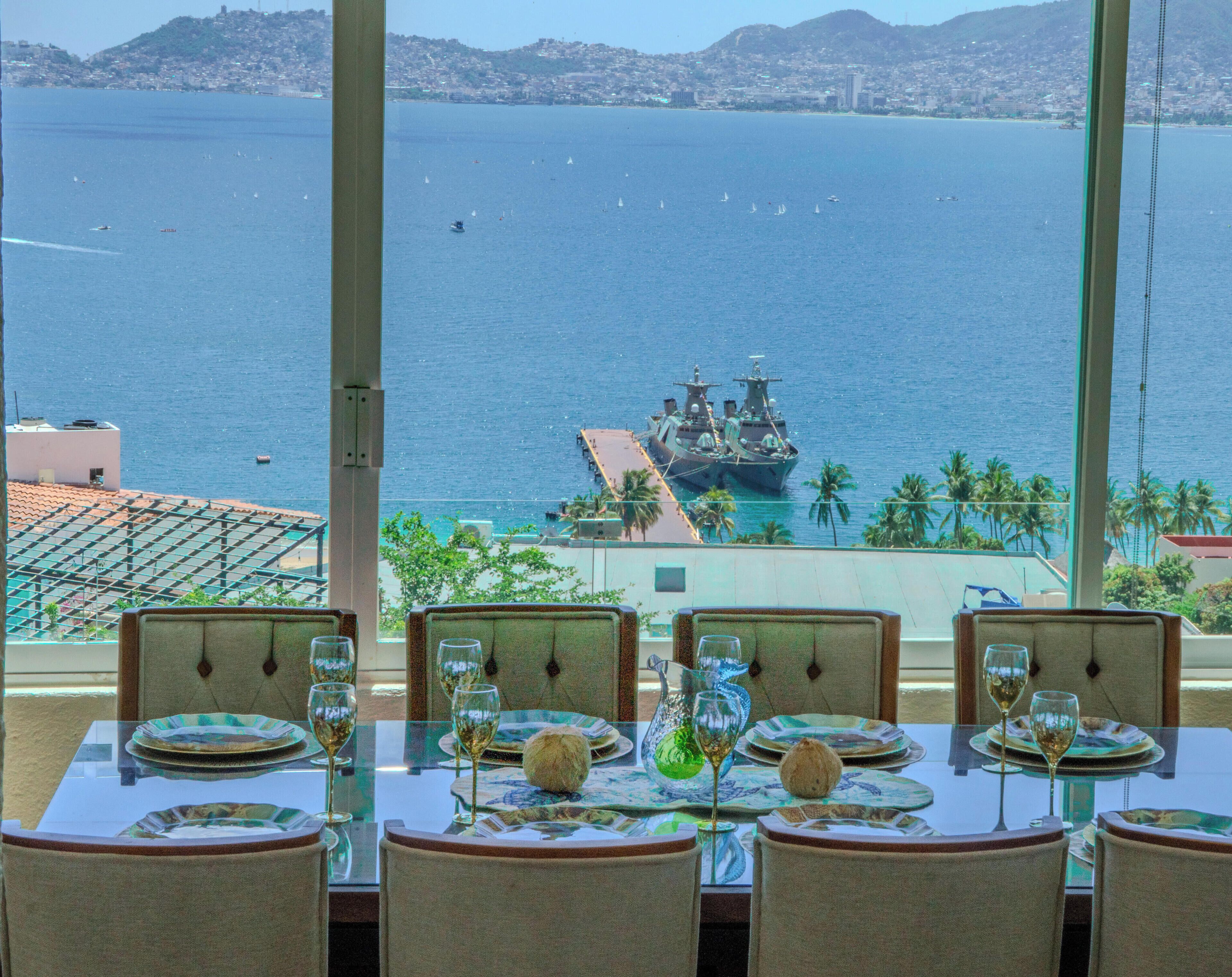 Mesa de comedor dispuesta con platos, vasos y un centro de mesa, con vistas a la costa, un barco y un paisaje urbano.