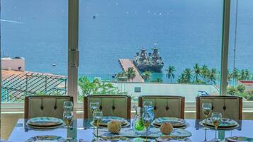 Mesa de comedor dispuesta con platos, vasos y un centro de mesa, con vistas a la costa, un barco y un paisaje urbano.