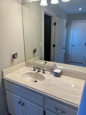 A bathroom with a large mirror, a sink with two faucets, a soap dispenser, and a small container. There are cabinets below the sink and a door in the background.