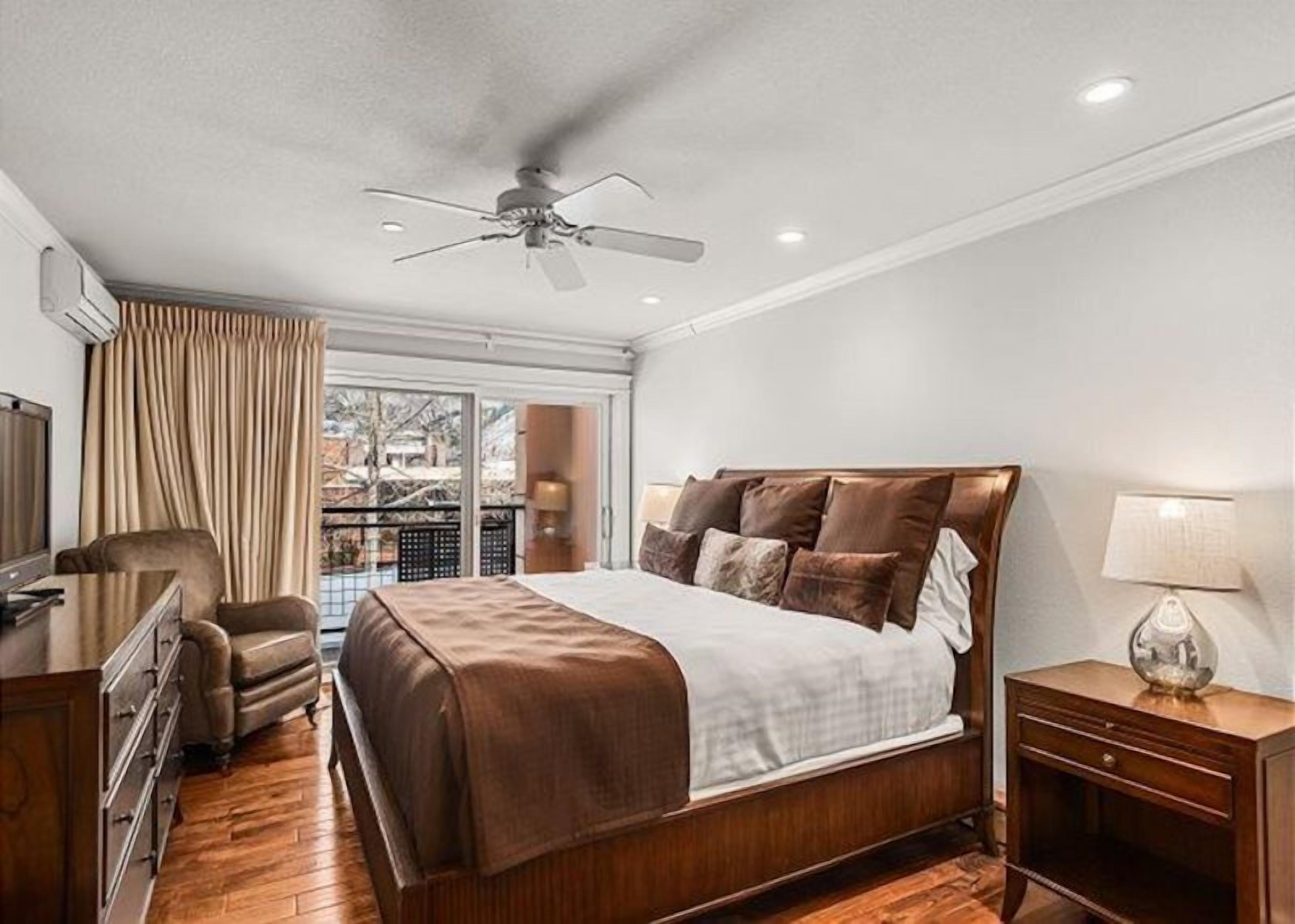 Habitación de hotel con una cama grande, ventilador de techo, televisor, una silla y ventana con cortinas.