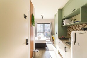 Cocina compacta con microondas, fregadero y refrigerador. Se ve una cama a través de una puerta abierta, y un balcón con vistas a edificios a través de una ventana.
