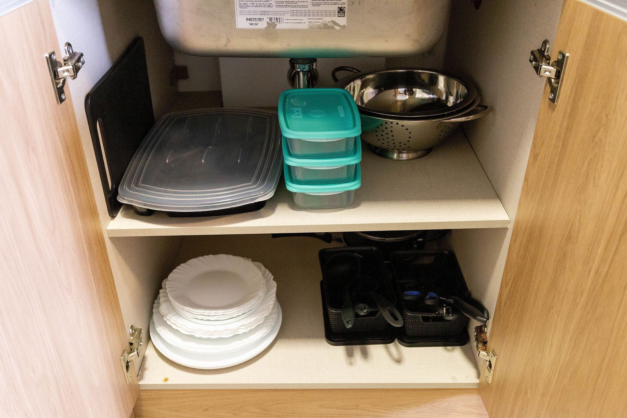 A kitchen cabinet with storage containers, a colander, and stacked plates.