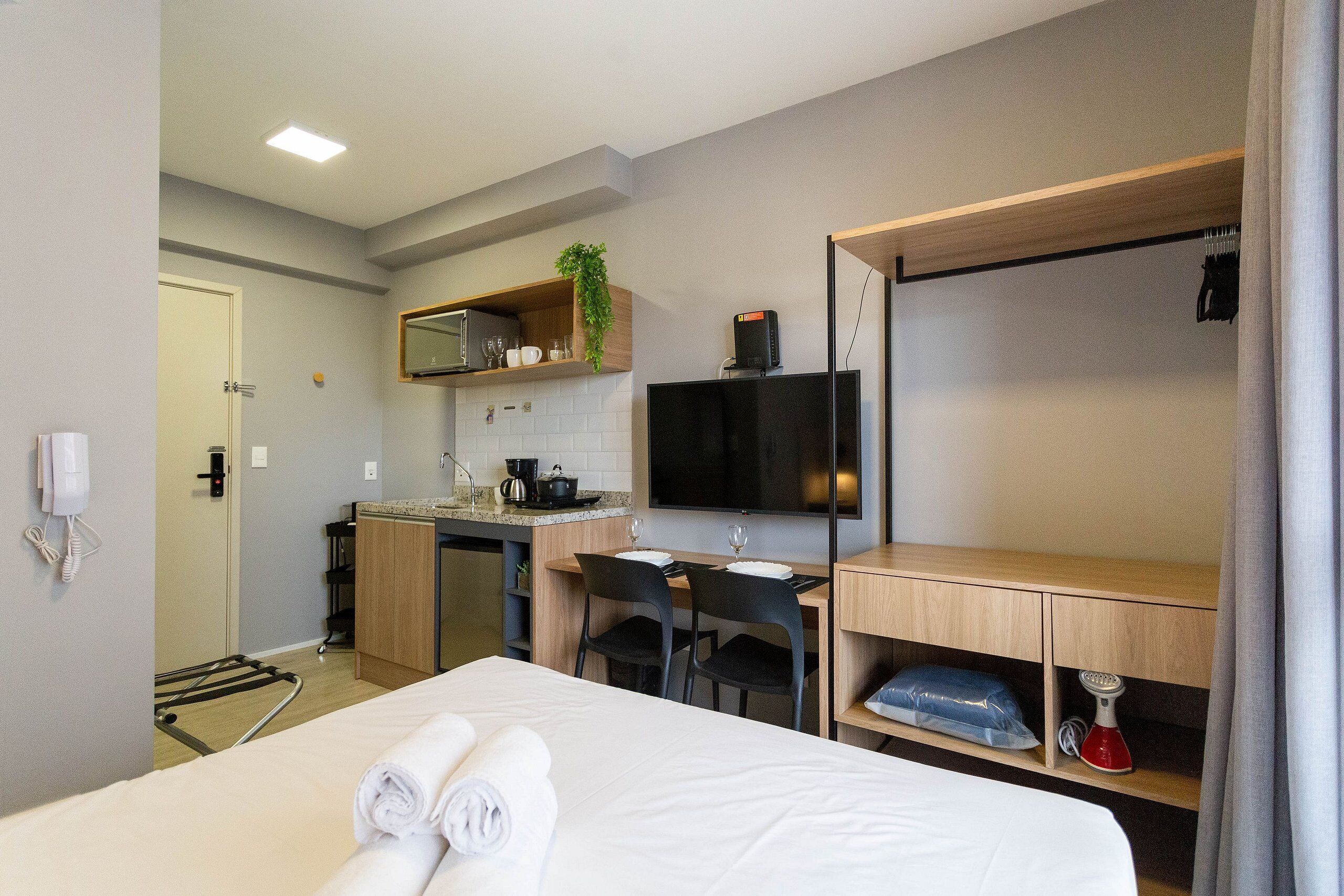 Ett hotellrum med en säng, ett skrivbord, en stol, en TV, en mikrovågsugn, ett handfat och en liten köksdel.