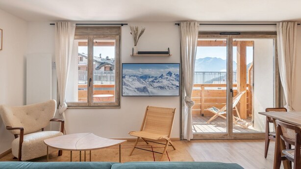 A cozy living room with a sofa, a small round table, and a chair. There is a balcony with a wooden deck and a view of mountains.